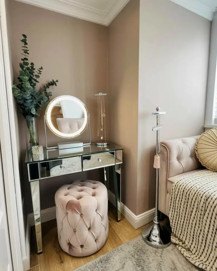 Mirrored vanity table with tufted ottoman in neutral bedroom corner. LED-lit round mirror and eucalyptus plant complete the setup.