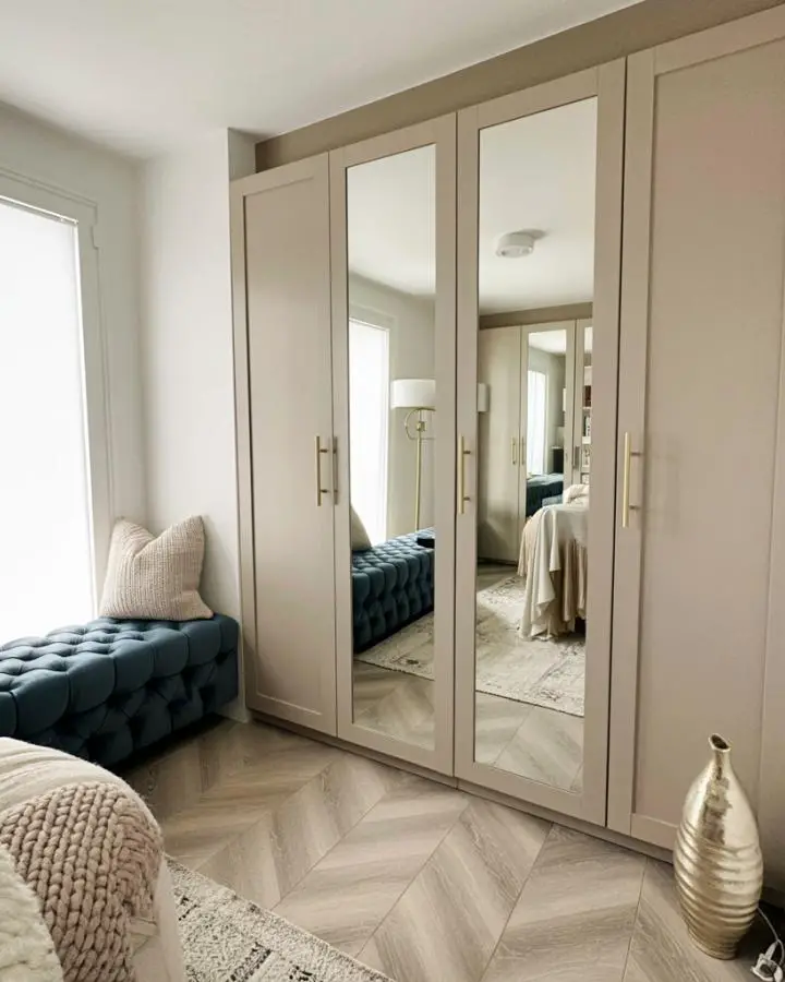 Beige built-in wardrobes with mirrored doors and navy tufted bench. Features herringbone flooring and natural lighting.