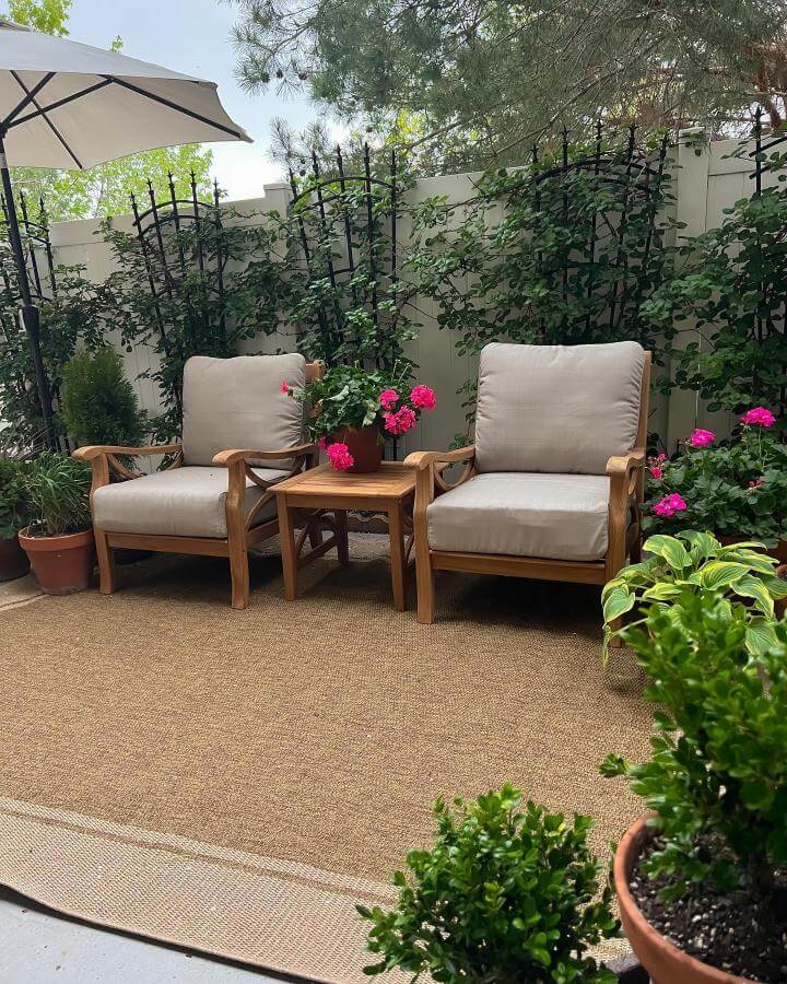 Beige cushioned wooden chairs with potted pink flowers and a woven rug create a snug outdoor seating area by @house.on.oak.lane. Lush plants surround the cozy space.