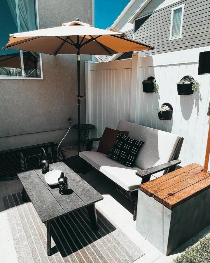 Neutral cushioned outdoor sofa with wood and concrete storage bench from @makingmycasita. Small space patio with vertical planters and beige umbrella shade.