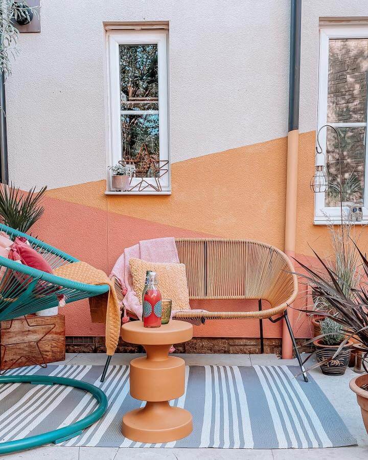 Teal woven chair and rattan loveseat on striped rug with peach and orange geometric painted wall from @my.interior.tales. Bright and cozy outdoor sitting corner.