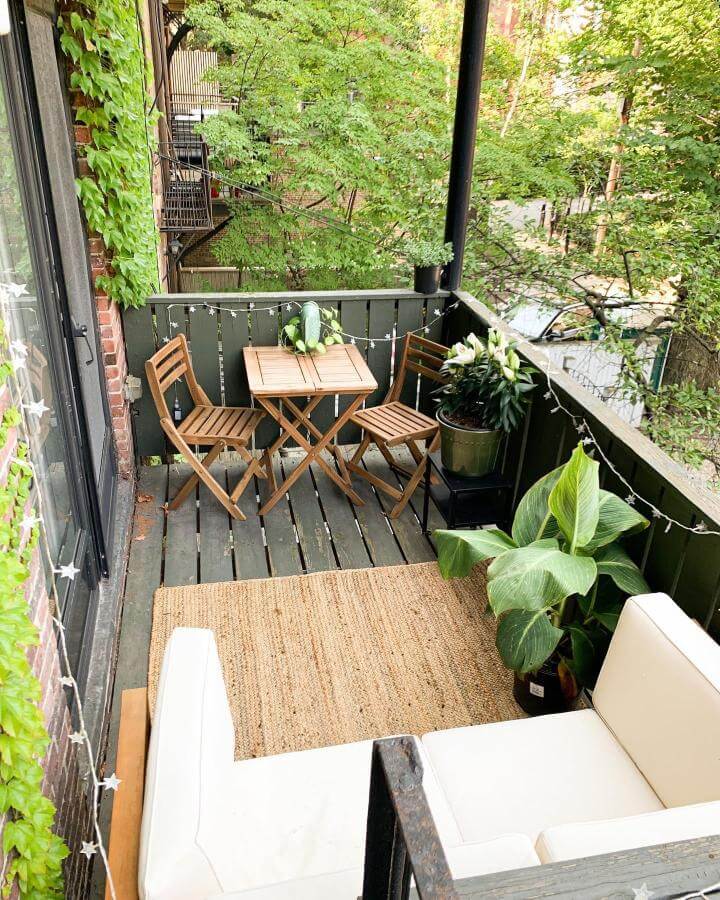 Natural wood bistro set and lush plants on cozy small outdoor balcony by @ewickdesign with inviting greenery and textured rug.