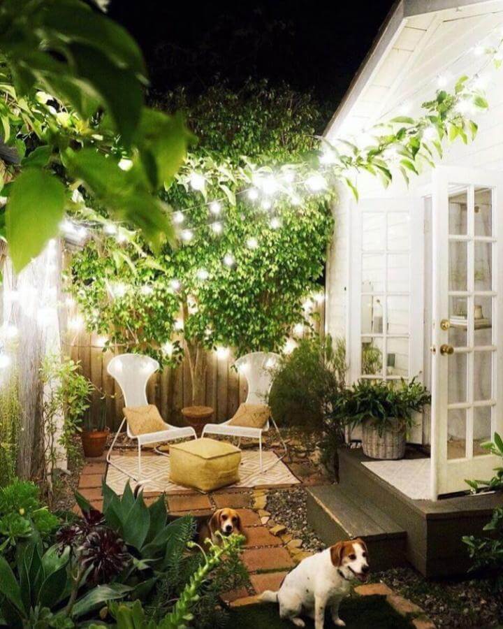 Warm string lights over white chairs and mustard pouf on patterned rug with lush greenery by @thekatrinablair creating an intimate backyard retreat.