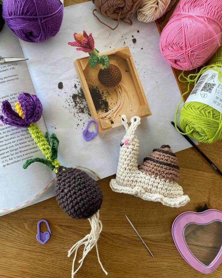 Crocheted spring snail and flowers in pastel pink, green, and brown yarn from @hobbii_yarn on wooden surface with yarn balls and crochet tools spring crafts for adults.