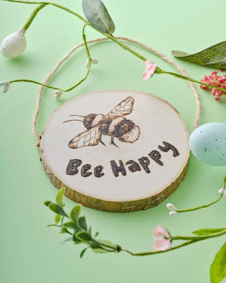 Natural wood slice with pyrography bee and cheerful phrase from @hobbycrafthq surrounded by spring crafts for adults decorative flowers and eggs.