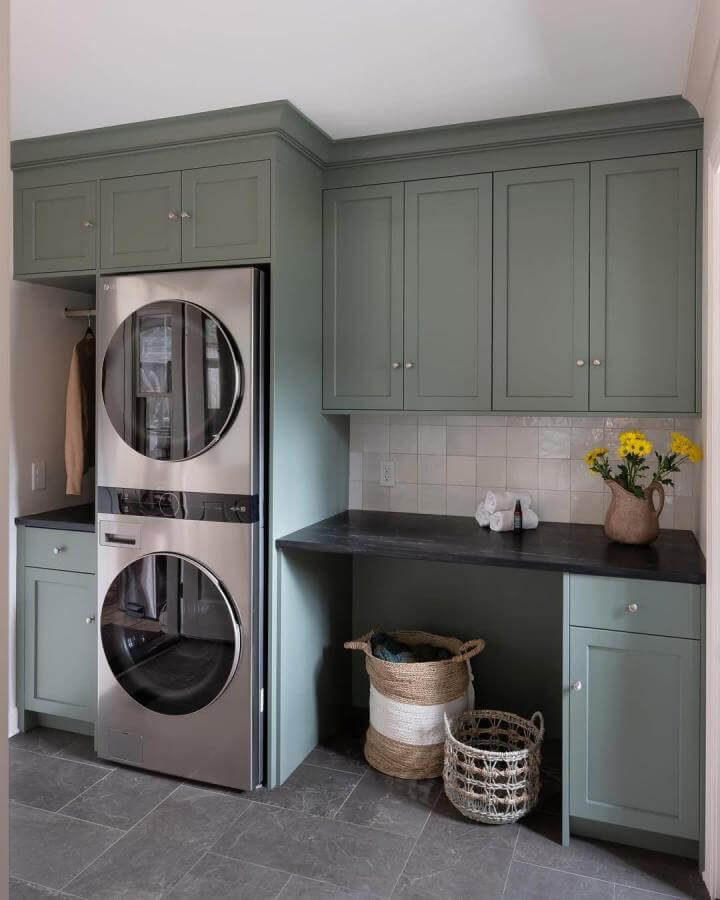 Muted sage cabinetry laundry room from @kitchensunlimited featuring stacked washer dryer laundry rooms with black countertops and woven baskets.