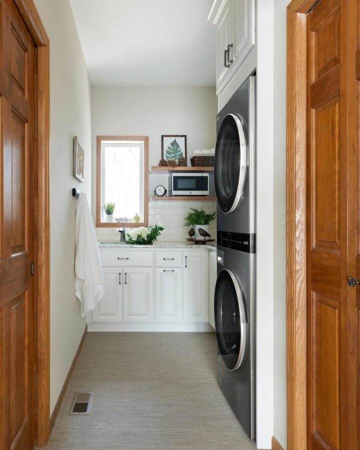 Bright white cabinet laundry space from @crystalkitchenbath with stacked silver units and warm wood trim around doors and window.