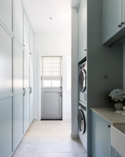 Soft blue shaker cabinetry laundry space from @strittdesignandconstruction with stacked washer and dryer and pale limestone flooring.