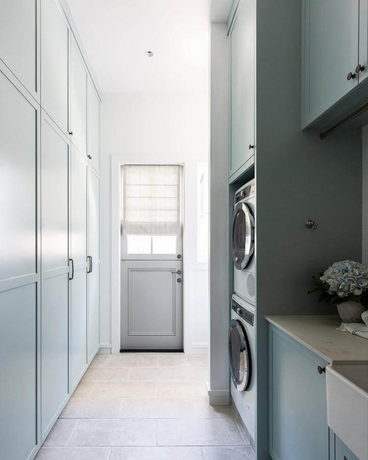 Soft blue shaker cabinetry laundry space from @strittdesignandconstruction with stacked washer and dryer and pale limestone flooring.