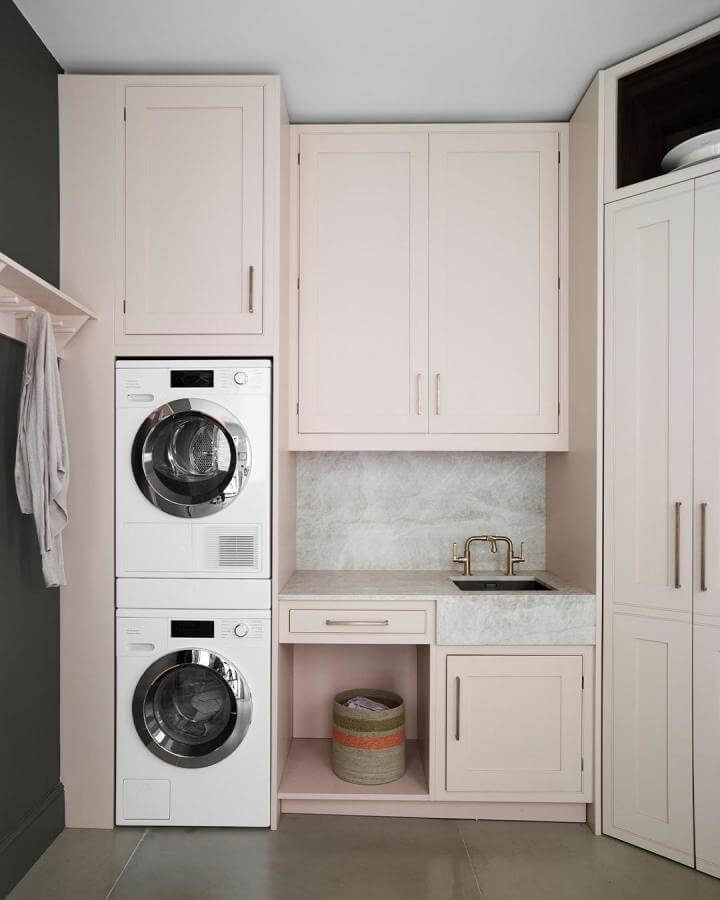 Soft blush cabinetry and marble-look surfaces in stacked washer dryer laundry rooms from @roundhouse_design with brass fixtures and cozy open shelving.