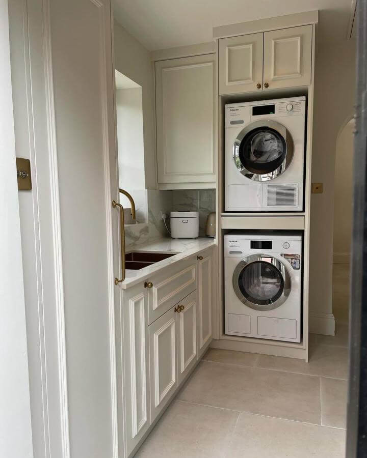 Cream cabinetry laundry space from @clarke_kitchens featuring brass handles and marble-effect countertop near stacked washer and dryer.