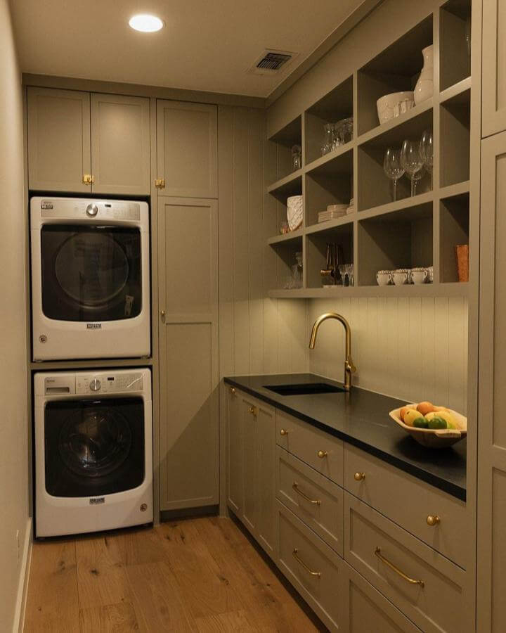 Olive green cabinetry laundry nook from @woodruffhomestx with stacked appliances black countertops and brass fixtures.
