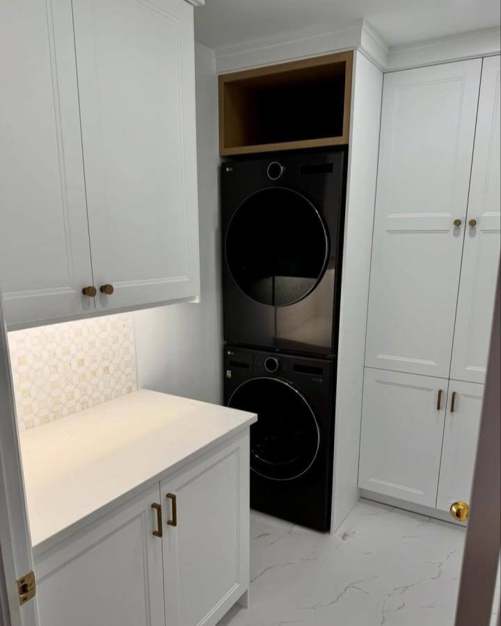 Sleek black stacked laundry machines with white cabinetry and brass hardware by @elliotcarpentry in a bright, organized laundry nook.