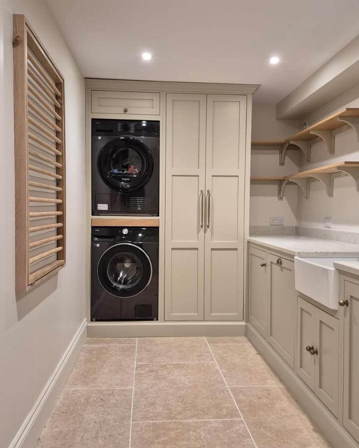 Neutral cabinetry laundry nook with black appliances and wooden shelves from @holland_kitchens. Compact and elegant utility space.