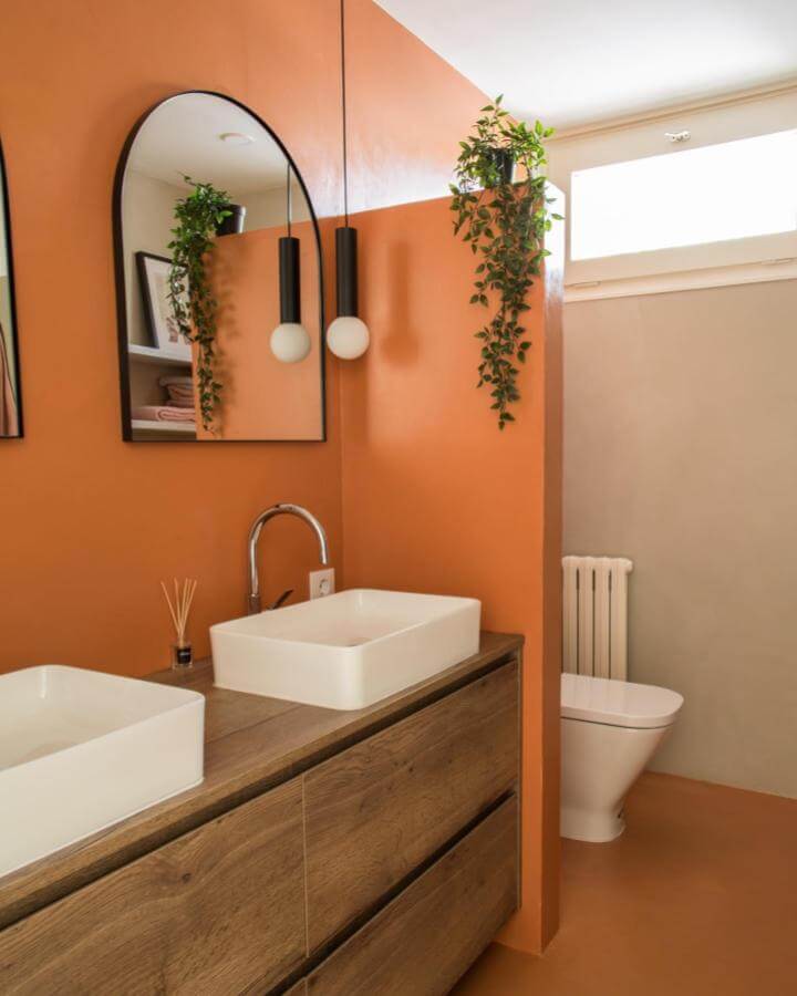 Warm terracotta bathroom walls with white sinks, wooden vanity, black pendant lights, and green hanging plants from @made_bcn_ creating cozy earthy vibe.