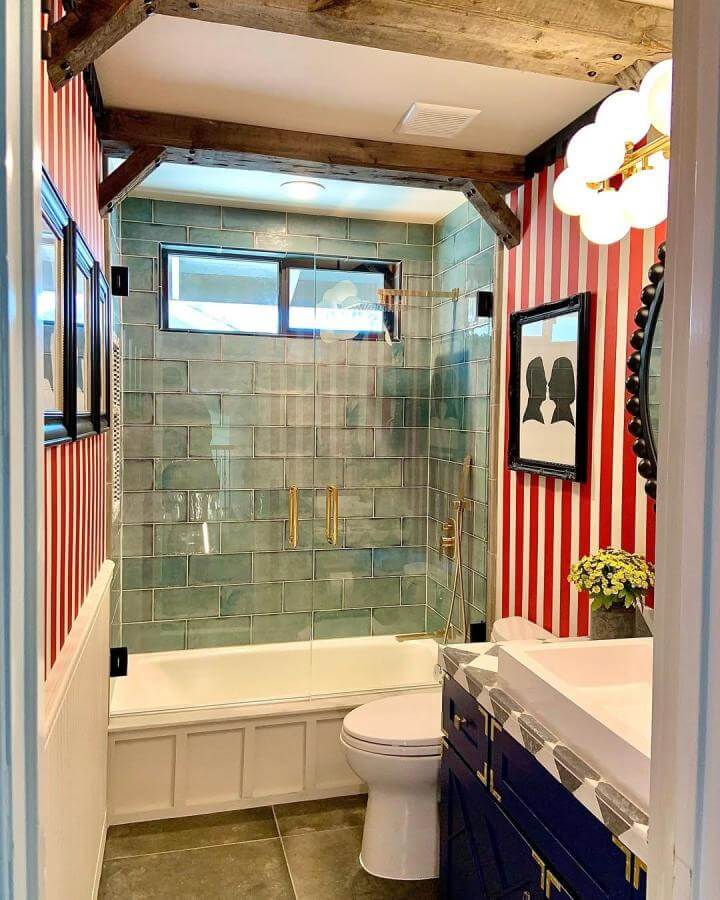 Blue-green glazed subway tile bathroom with brass hardware and wood beams from @oldeenglishtiles. Small space with vibrant striped wallpaper and clear glass shower.