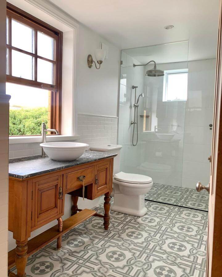 Wooden vintage vanity with marble countertop and patterned encaustic floor tiles in a Victorian bathroom ideas setting by @biancasantosdesigns bright natural light window and glass shower.