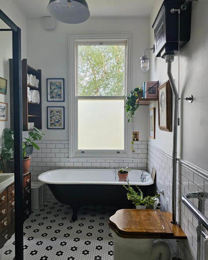 Black clawfoot bathtub with white interior, hex tile floral floor, and wood accents in a Victorian bathroom from @homeisheathside. Bright window and wall art enhance charm.