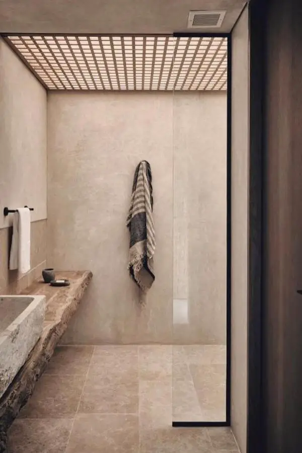 Wooden slatted ceiling bathroom from @ancre.home. Natural light creates geometric shadow patterns on beige stone walls.
