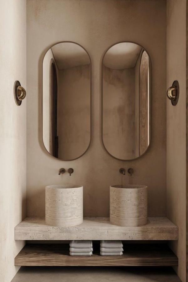Warm beige bathroom vanity featuring dual oval mirrors and textured stone vessel sinks. Minimalist design with natural materials and brass accents.