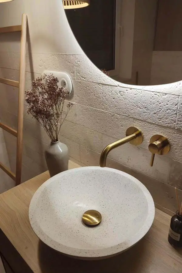 Textured cream stone bathroom tiles with brass fixtures and natural wood vanity. Rough surface creates beautiful shadows and organic texture.