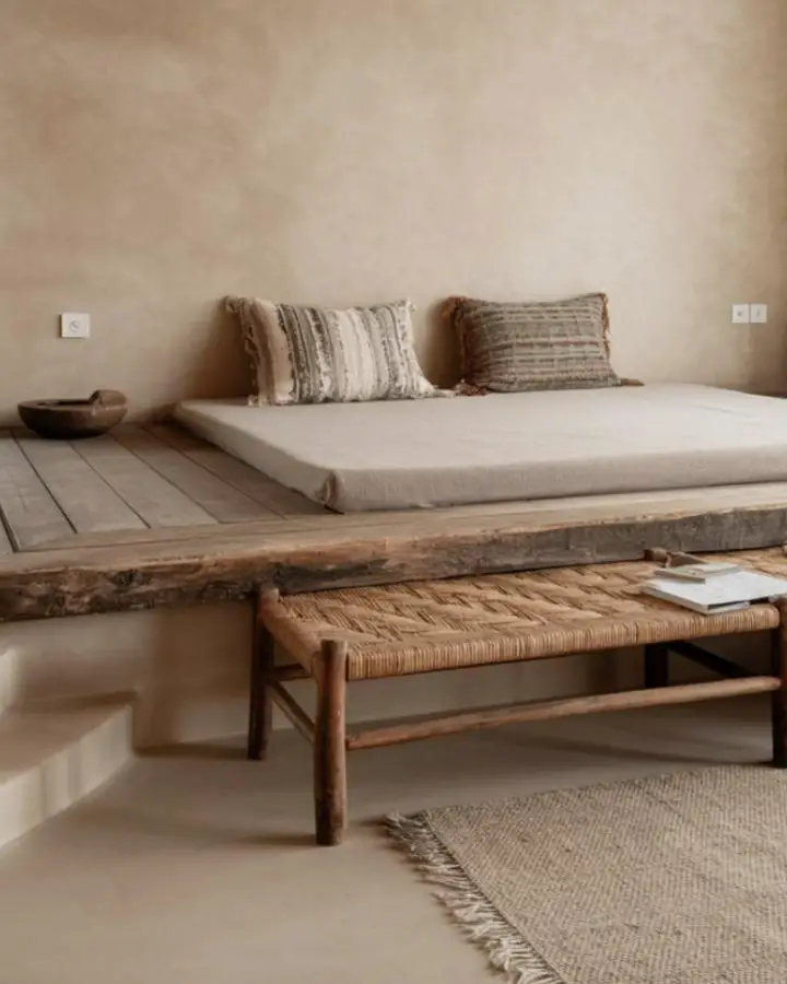 Weathered wood platform bed with woven bench in neutral bedroom featuring natural textures and cream bedding.