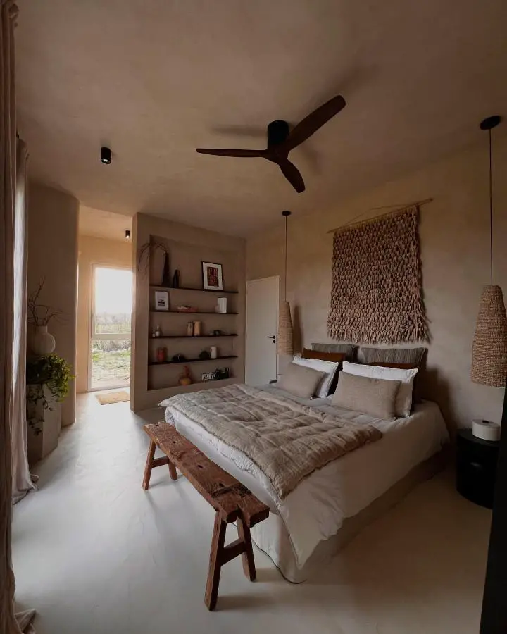 Neutral bedroom from @maisontoscane featuring handwoven macrame wall art above wooden bench and linen bedding.