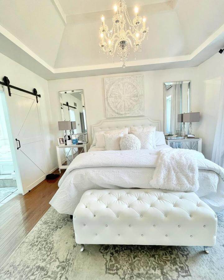 White layered bedroom with tufted bench and chandelier from @thealexandersnest featuring cozy textures and reflective accents.