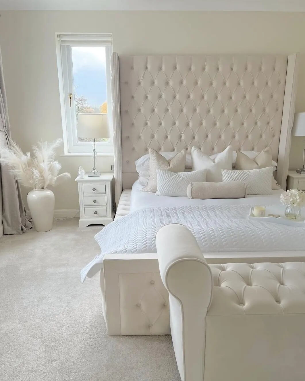 Creamy tufted upholstered bed and bench from @puresaltinteriors with soft linens and pampas grass accents. Bright and serene bedroom setting.
