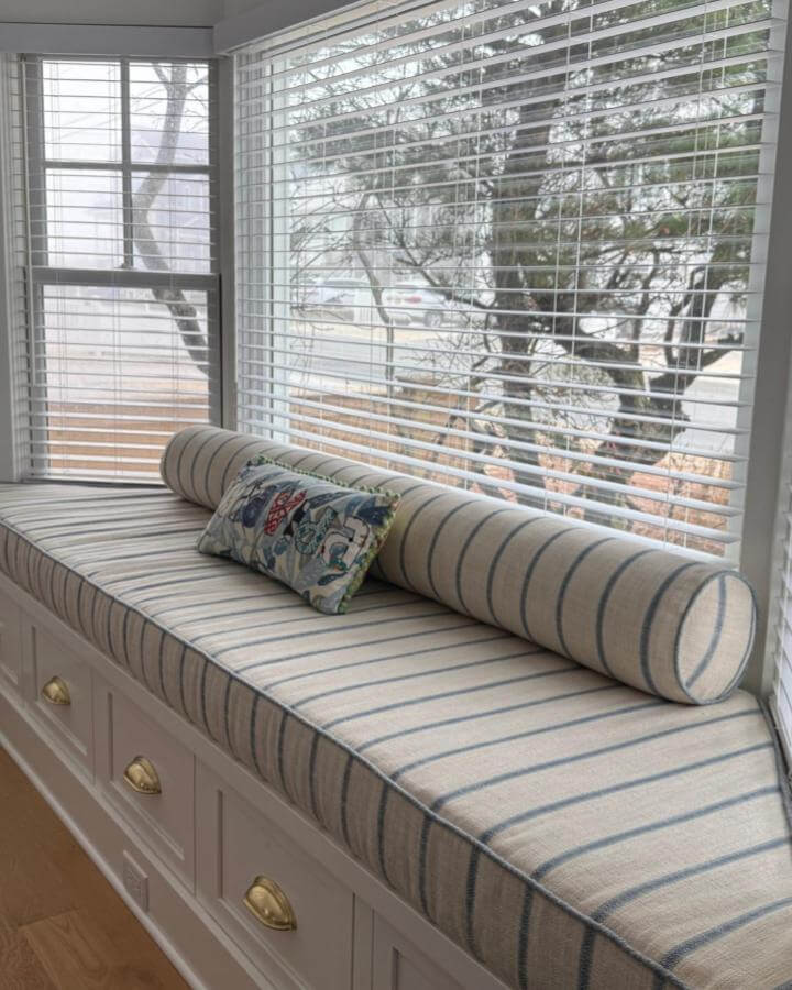 Light blue striped cushioned window bench with built-in white drawers from @curated_by_ellen in a bright cozy room.