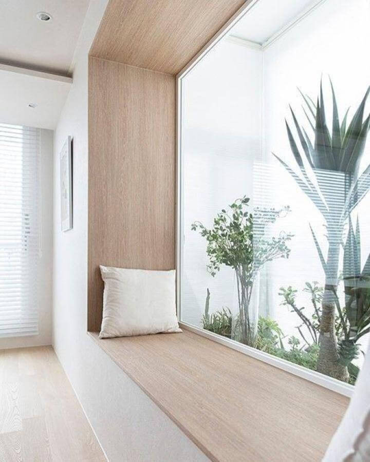 Light oak wood window nook seating from @alenacdesign featuring minimalist cushion and large glass overlooking greenery.