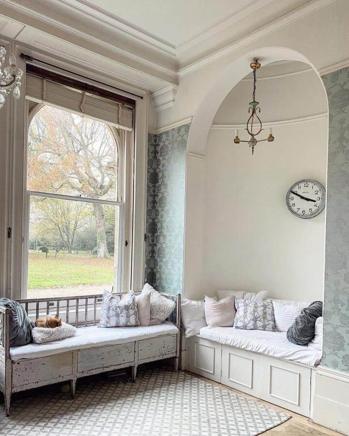 White distressed wooden window seat with soft cushions and storage from @thevintagetraderuk in a bright, vintage-inspired interior.