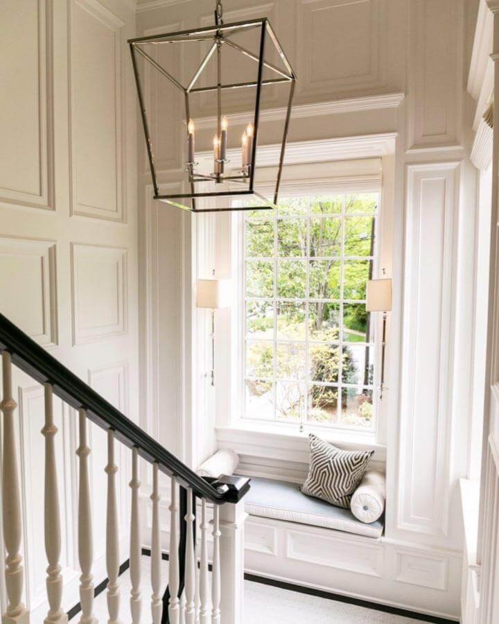 Light gray cushioned window nook with white paneled walls and black metal lantern pendant light from @sandyspringbuilders. Quiet, elegant stairway reading corner.