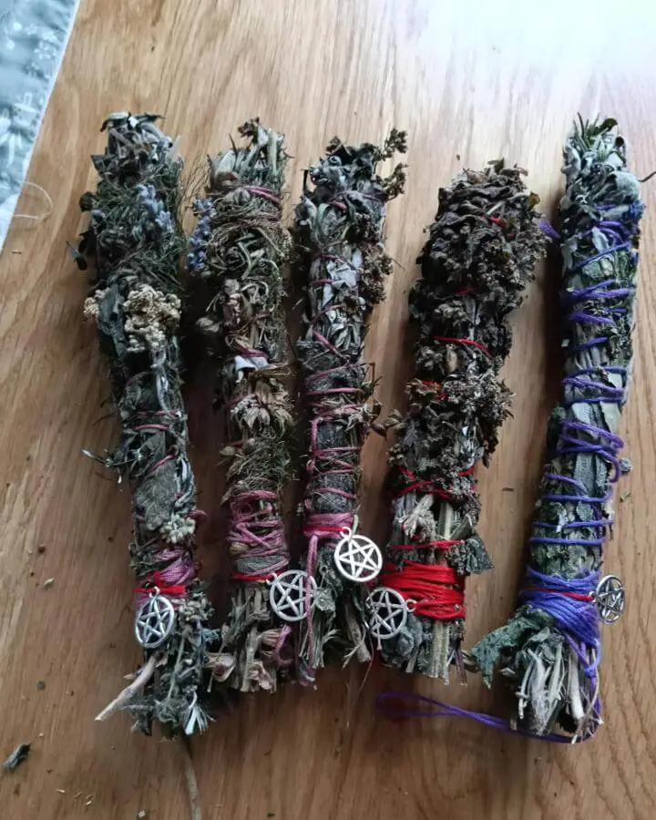 Handmade herb bundles from @samgreenwitch arranged on wooden surface with colorful thread wrapping and pentacle charms attached.