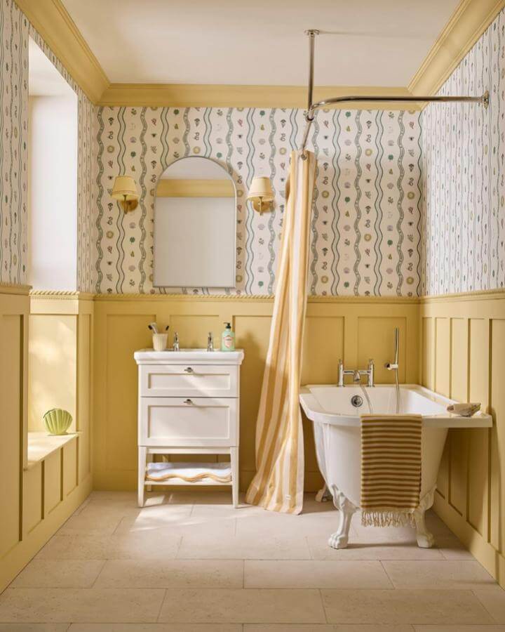 Warm yellow wainscoting and striped accents in a bright bathroom from @orac_uk with vintage clawfoot tub and patterned wallpaper.